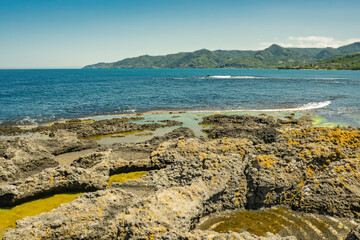 Scenic coastal landscape featuring rugged volcanic rocks with yellow lichen. Shallow tidal pools, and the deep blue sea extending toward distant green mountains under blue sky. Turkey, Black sea.