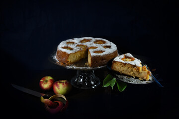 Moist, fresh, homemade apple cake baked according to a traditional German recipe. Sponge cake with apples and powdered sugar. Dark surroundings, studio photography.