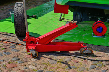 Red hydraulic floor jack for lifting cars during maintenance or tire replacement. Professional garage equipment on cobblestone pavement, concept of auto repair, service, and workshop tools.
