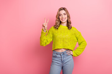 Obraz na płótnie Canvas Cheerful young woman in stylish yellow knit sweater giving a peace sign gesture, smiling against a pink background