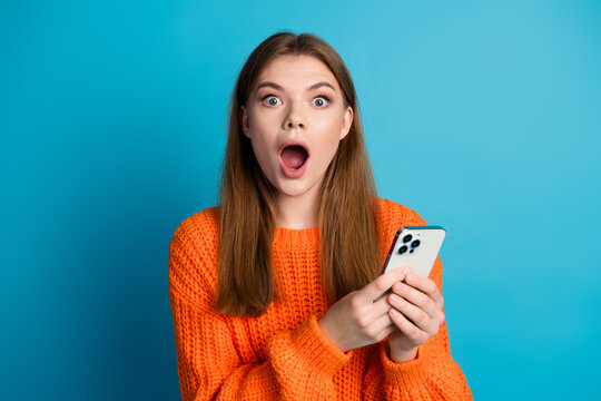 Young woman in orange sweater looks amazed while holding smartphone, posing against blue background