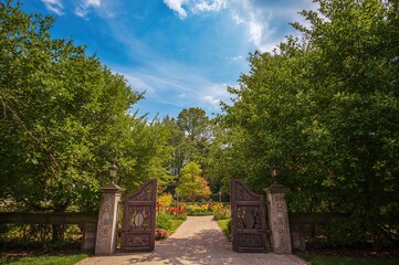 Scenic garden gateway under a bright sky with fluffy clouds, surrounded by vibrant plants and numerous trees forming a peaceful setting.