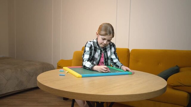 Primary school student practicing drawing on a small blackboard placed on a round wooden table near a mustard-colored sofa in a cozy living room in an apartment