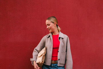 Young woman with paper parcel by a red wall opens online order pickup. Contactless delivery, eco packaging, click&collect, last-mile logistics, D2C retail, unboxing, customer, smile urban lifestyle