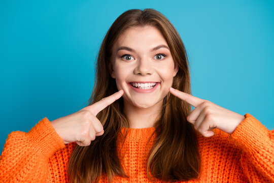 Joyful young woman pointing to her bright smile wearing an orange sweater against a vibrant blue background
