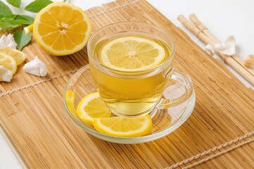 Transparent glass cup filled with lemon-infused tea, placed on a bamboo mat with lemon slices on a white surface.