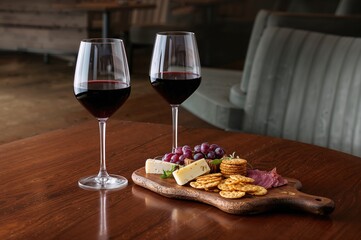 Naklejka premium Two glasses of red wine accompanied by a wooden board with grapes, cheese, and crackers on a dining table. Concept for wine enthusiasts.