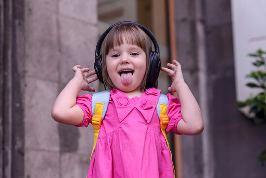 Cheerful little girl in pink dress with backpack listening to music in big headphones in front of stone wall. Laughing child showing tongue and enjoying life. Concept of childhood, freedom, and joy.
