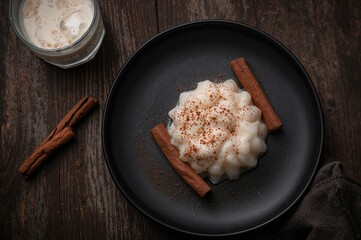 A single portion of creamy rice pudding garnished with powdered cinnamon and cinnamon sticks, served on a dark plate next to a glass of milk on a wooden surface.
