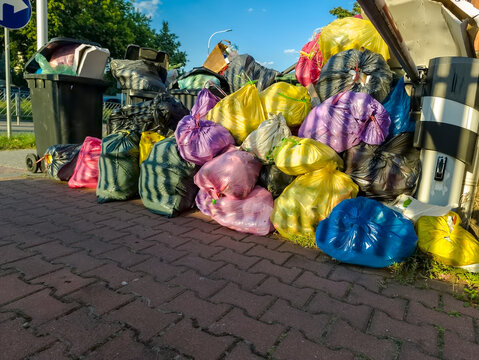 Malowniczo kolorowy stos śmieci w workach i koszach na śmieci w słońcu letniego popołudnia. Stos kolorowych work&oacute;w na śmieci, wysypujący się na chodnik. Worki są w odcieniach ż&oacute;łci, r&oacute;żu, błękitu 