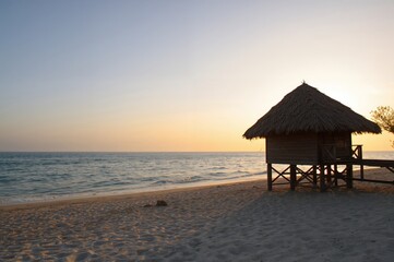 A peaceful seaside scene with a wooden cabin elevated on stilts by the shore, overlooking calm waters bathed in the glow of a setting sun.