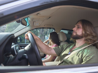 Naklejka premium Casual car interior scene featuring a man driving with both hands on the wheel while a passenger sits in the back, soft daylight creating a relaxed mood.