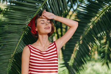 A cheerful woman enjoys a sunny moment outdoors among lush palm fronds, wearing a red and white striped top. She smiles softly with eyes closed, savoring warm sunlight.