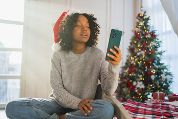 Merry Christmas. African woman in Santa hat having video chat in smartphone. Blogger in festive mood recording video in social network. Girl having video call with friends family celebrating Christmas