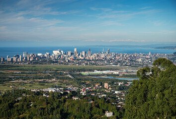 Obraz premium Aerial view of modern coastal city with buildings, green fields and trees in foreground, mountains