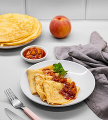 Pancakes with peach jam on a plate on a light kitchen brick background with fresh fruits, napkin and cutlery.