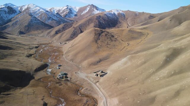 Wide aerial view of Tash Rabat caravanserai surrounded by mountain landscape, river, and snowy peaks. A historic Silk Road monument located in the Tien Shan mountains.