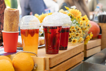 Plastic cups with berry and citrus drinks, traditional pastries and fresh oranges on a rustic wooden counter at a local fair, with flowers and people in the background