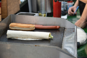 Close-up of a hot grill at a street food stand with a tortilla wrap, sausage, and bun being heated, typical fast food preparation at outdoor market