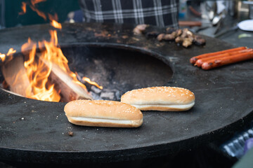 Hot dog buns and sausages being grilled on a round open fire grill at a street food event, capturing the dynamic and rustic feel of outdoor cooking.