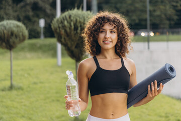 Sportswoman holding water bottle and yoga mat, smiling in a park