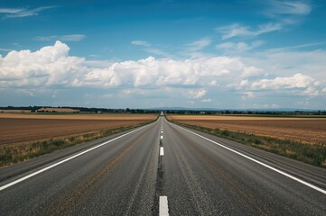 Fototapeta premium A long paved highway crossing farmland beneath a bright sky dotted with soft white clouds, extending far ahead. Theme of automotive journey and exploration.