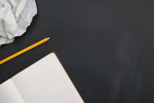 Bright yellow pencil resting on a notebook beside wrinkled sheets on a blackboard background - Powered by Adobe