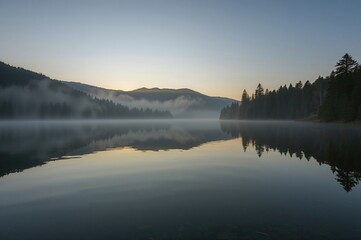Fototapeta premium A fog-covered lake mirrors the outline of gentle hills under the warm early morning light, forming a calm and harmonious natural landscape.