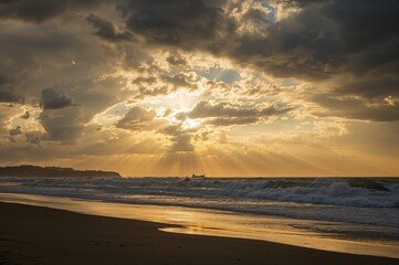 Obraz premium Coastal scene under a turbulent sky with sunlight breaking through, sandy shore at dusk featuring ocean waves and darkened landforms.