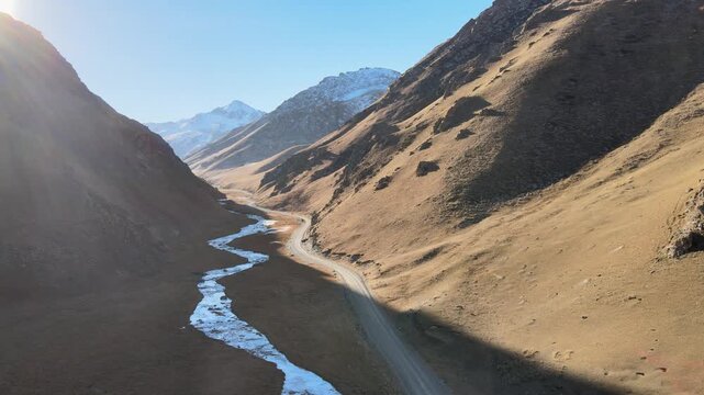 Aerial view of a mountain road running through a valley along a river, surrounded by mountains with snowy peaks. The road passes at an altitude of over 3500 meters above sea level and leads to the his