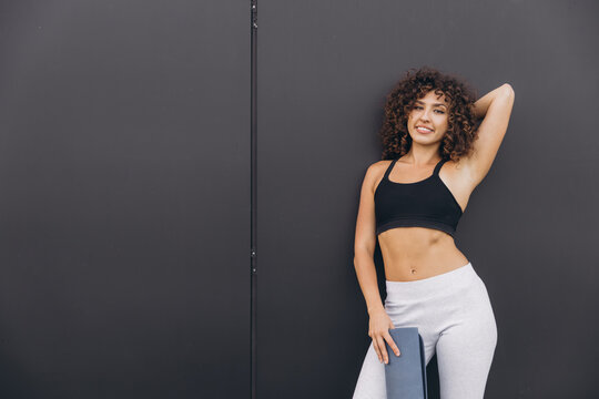 Young fit woman holding yoga mat posing against black wall