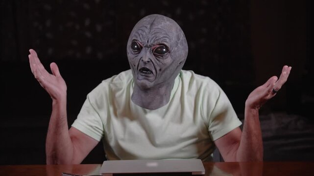 A person in a realistic alien mask sits at a table with arms raised in a shrugging gesture, expressing confusion, disbelief, or frustration in a dramatic pose. 14827  	