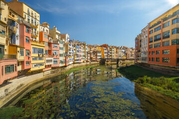 Colorful houses mirrored in the calm river Onyar, Girona’s Barri Vell under blue summer sky, Florentine feel; ideal for travel and city branding.