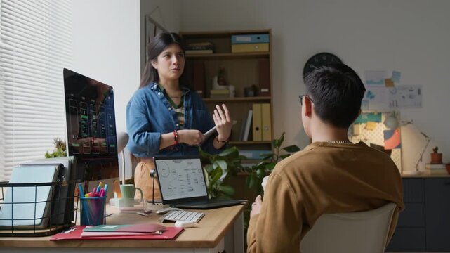 Back view of unrecognizable male programmer taking sip of coffee while listening to talkative female colleague who standing in front of him holding tablet