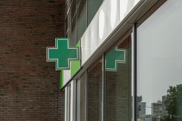 green cross, a common symbol for a pharmacy, is mounted on the corner of a brick building. The sign is reflected in the large glass windows of the modern facade