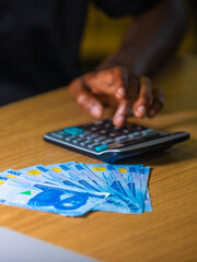 Dark skinned African person sitting punching pressing calculator with one thousand Nigerian naira notes on table desk