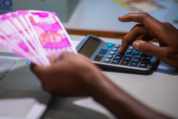 Dark skinned African person sitting punching pressing calculator with Indian Rupee notes on table desk