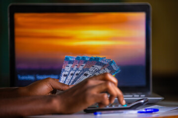 Dark skinned African person working on desk holding Saudi riyal notes in hand and pressing punching calculator with blue pen  and laptop on table. accounting concept.