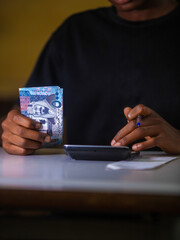 Dark skinned African person working on desk holding Saudi riyal notes in hand and blue pen pressing punching calculator on table. accounting concept.