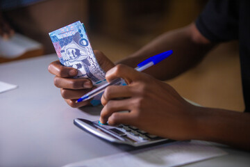 Dark skinned African person working on desk holding Saudi riyal notes in hand and pen pressing punching calculator  on table. accounting concept.