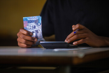 Dark skinned African person working on desk holding Saudi riyal notes in hand and blue pen pressing punching calculator on table. accounting concept.