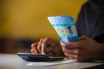 Dark skinned African person in black top working on desk holding Kuwaiti Dinar notes in hand and pressing punching calculator with receipt on table. accounting concept. 
