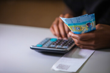 Dark skinned African person in black top working on desk holding Kuwaiti Dinar notes in hand and pressing punching calculator with receipt on table. accounting concept.