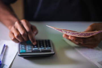 Top view of Dark skinned African person working on white desk pressing punching calculator and holding Ghanaian cedi notes in hand with more money on desk. accounting concept