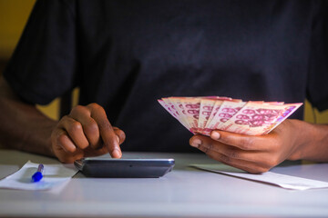 Dark skinned African person in black shirt working on white desk pressing punching calculator and holding Ghanaian cedi notes in hand. Accounting concept