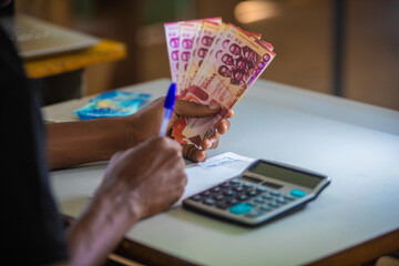 Dark skinned African person working on desk writing in notebook and holding Ghanaian cedi notes in hand. accounting concept