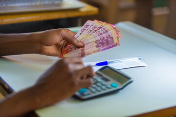 Dark skinned African person working on white desk pressing punching calculator and holding Ghanaian cedi notes in hand. Top view Accounting concept