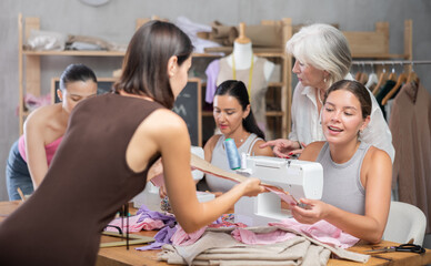 In sewing class for group of beginners, enthusiastic young woman working on machine, stitching fabric pieces of garment and discussing pattern with classmate..