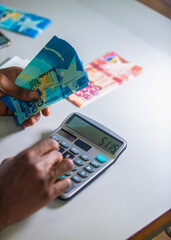 Dark skinned African person working on desk holding Ghanaian cedi notes in hand and pressing punching calculator with some notes on table. Top view accounting concept
