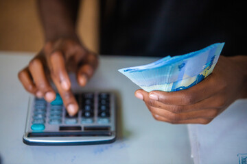 Dark skinned African person working on desk holding Ghanaian cedi notes in hand pressing punching calculator. accounting concept